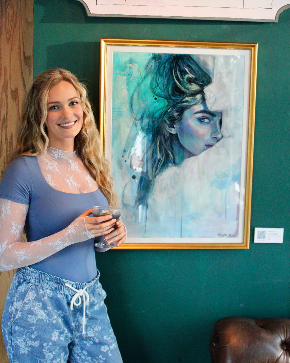 A woman stands next to a large framed portrait painting of a woman with teal and dark blue hair, set against a dark green wall.