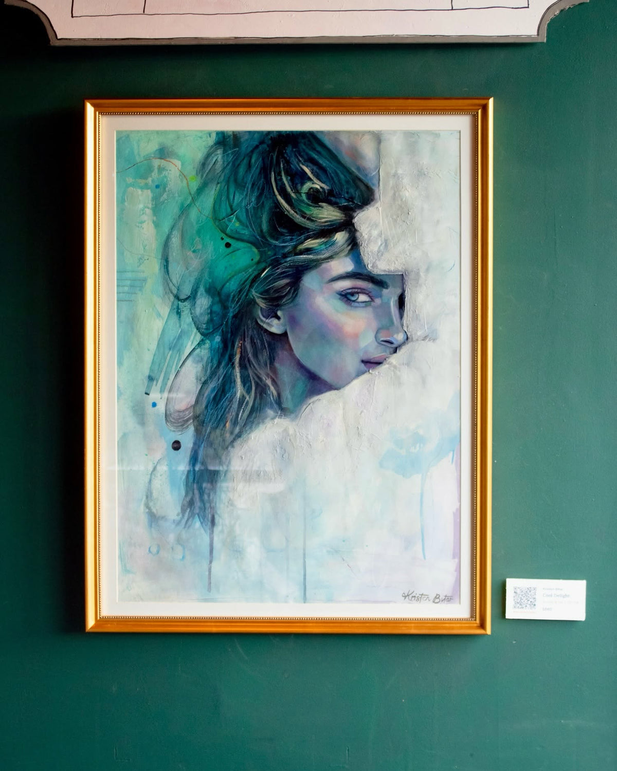 A framed portrait painting of a woman with blue, teal, and white tones, displayed on a dark green wall. The artwork is enclosed in a gold frame, with a small label card visible to the right.