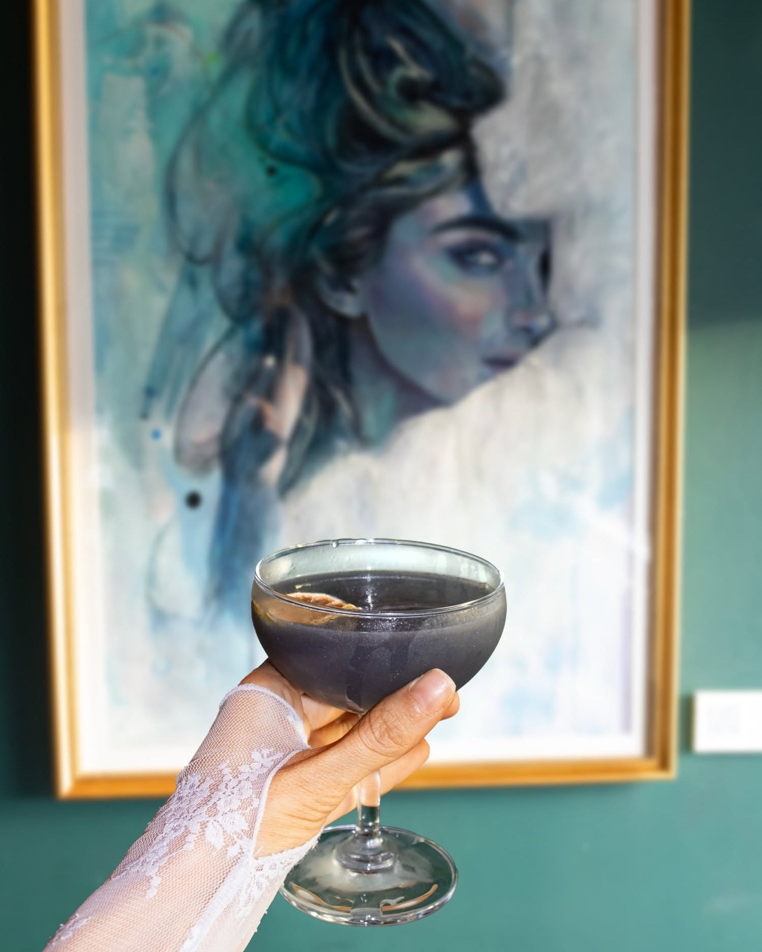 A hand in a white lace sleeve holds a coupe glass filled with a dark purple liquid in front of a framed portrait painting on a teal wall.