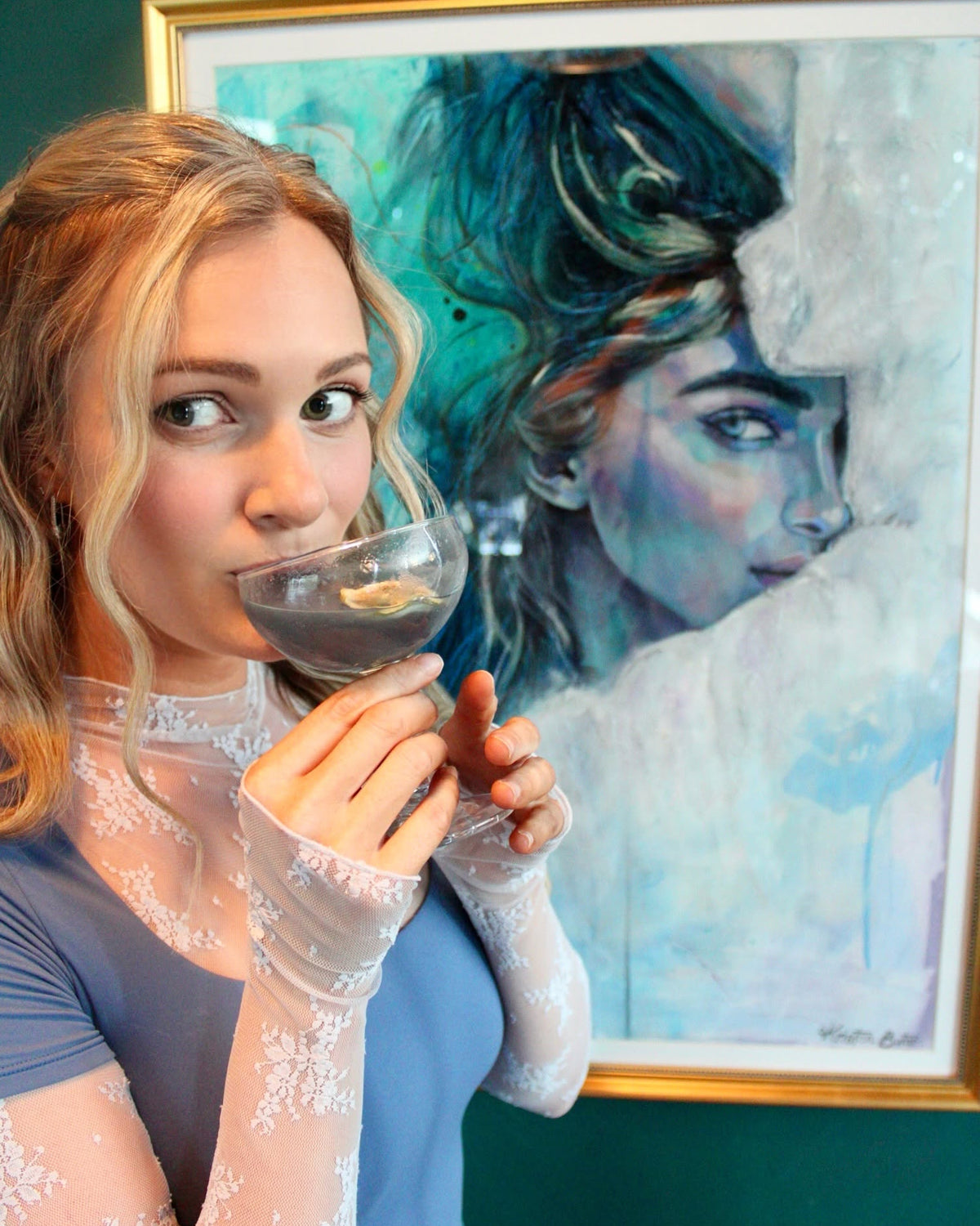 A woman in a blue t-shirt and sheer lace gloves holds a cocktail glass in front of a framed portrait painting of a woman with teal and white tones.