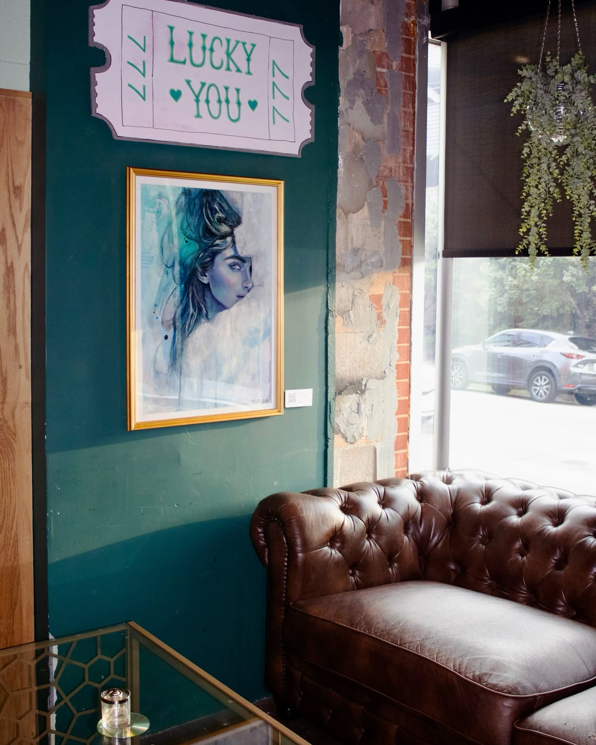 A styled interior corner featuring a dark brown tufted leather sofa next to a window, with a dark teal wall decorated with a 'LUCKY YOU' sign and a framed portrait of a woman.